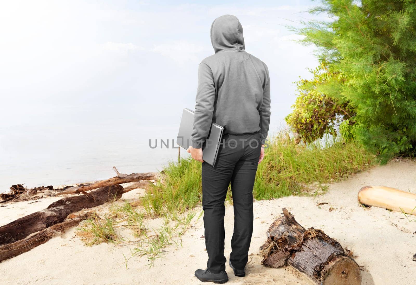 Rear view of man in black hoodie carrying laptop in his hand on the beach