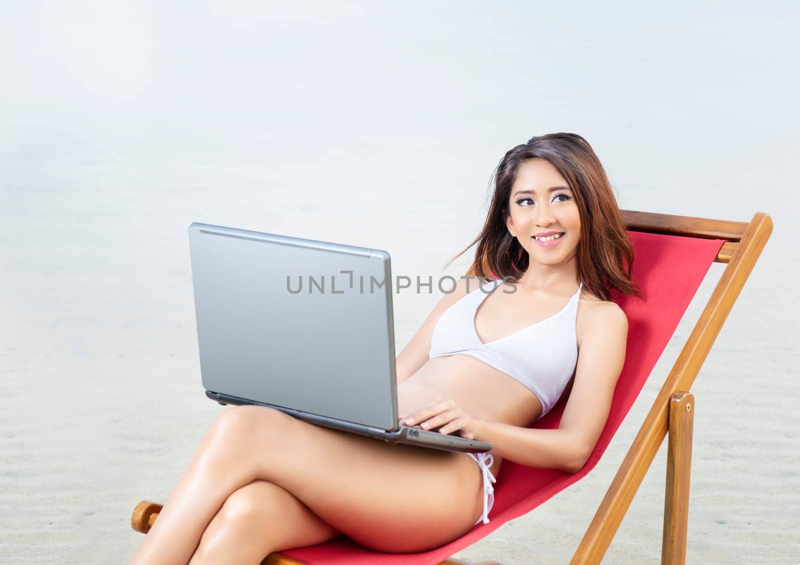 Asian sexy girl in bikini with laptop sitting on the beach chair at seaside. Summer vacation