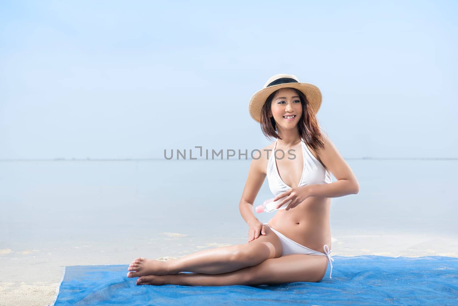 Asian sexy girl in bikini and hat sitting on the carpet and applying oil onto skin in beach. Summer vacation