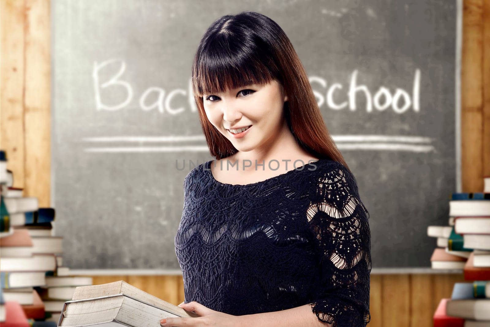 Asian student woman holding books on her hands in the classroom with piles of books and blackboard background. Back to School concept