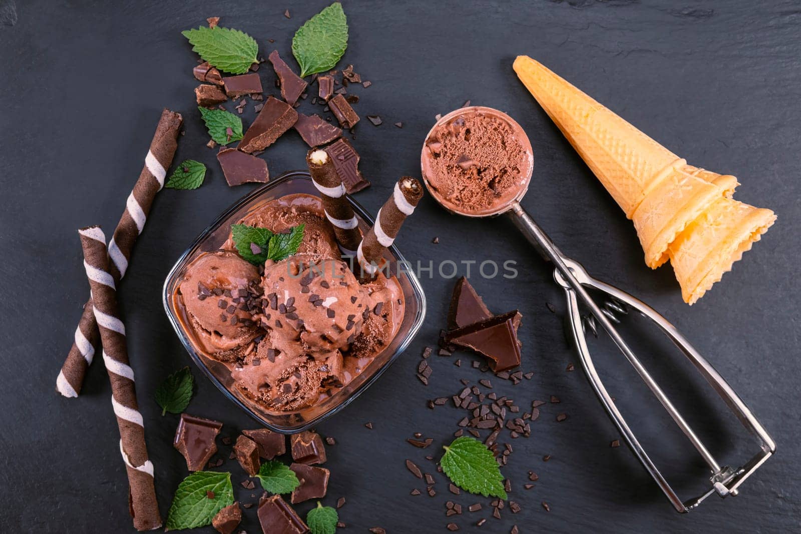 Scoops chocolate ice cream in glass bowl with wafer sticks, cone and chocolate on a black slate board. Focus on Bowl with scoops chocolate ice cream