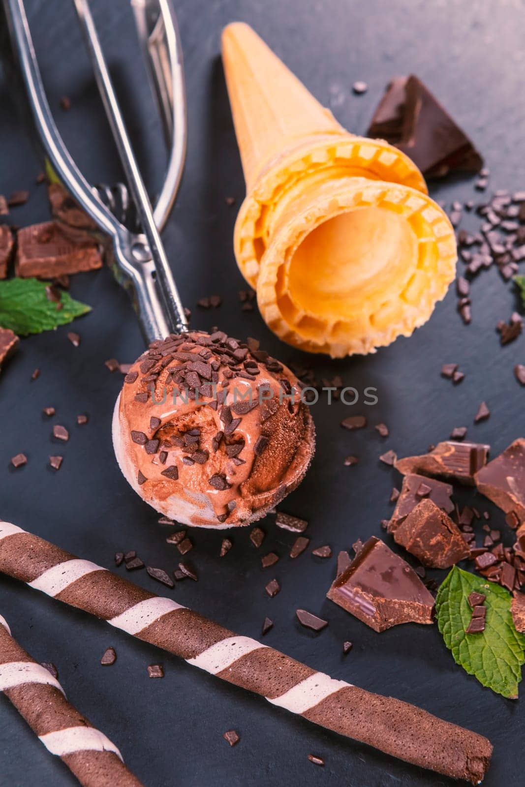 Chocolate ice cream in scoop with wafer sticks, cone and chocolate on a black slate board. Focus on ice cream in scoop.