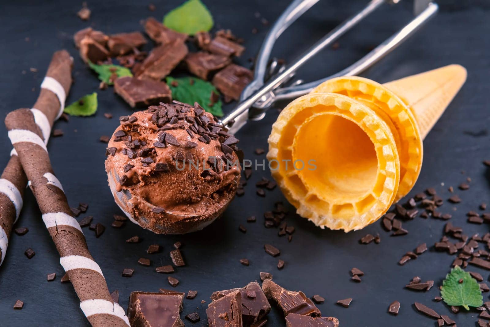 Chocolate ice cream in scoop with wafer sticks, cone and chocolate on a black slate board. Focus on ice cream in scoop.