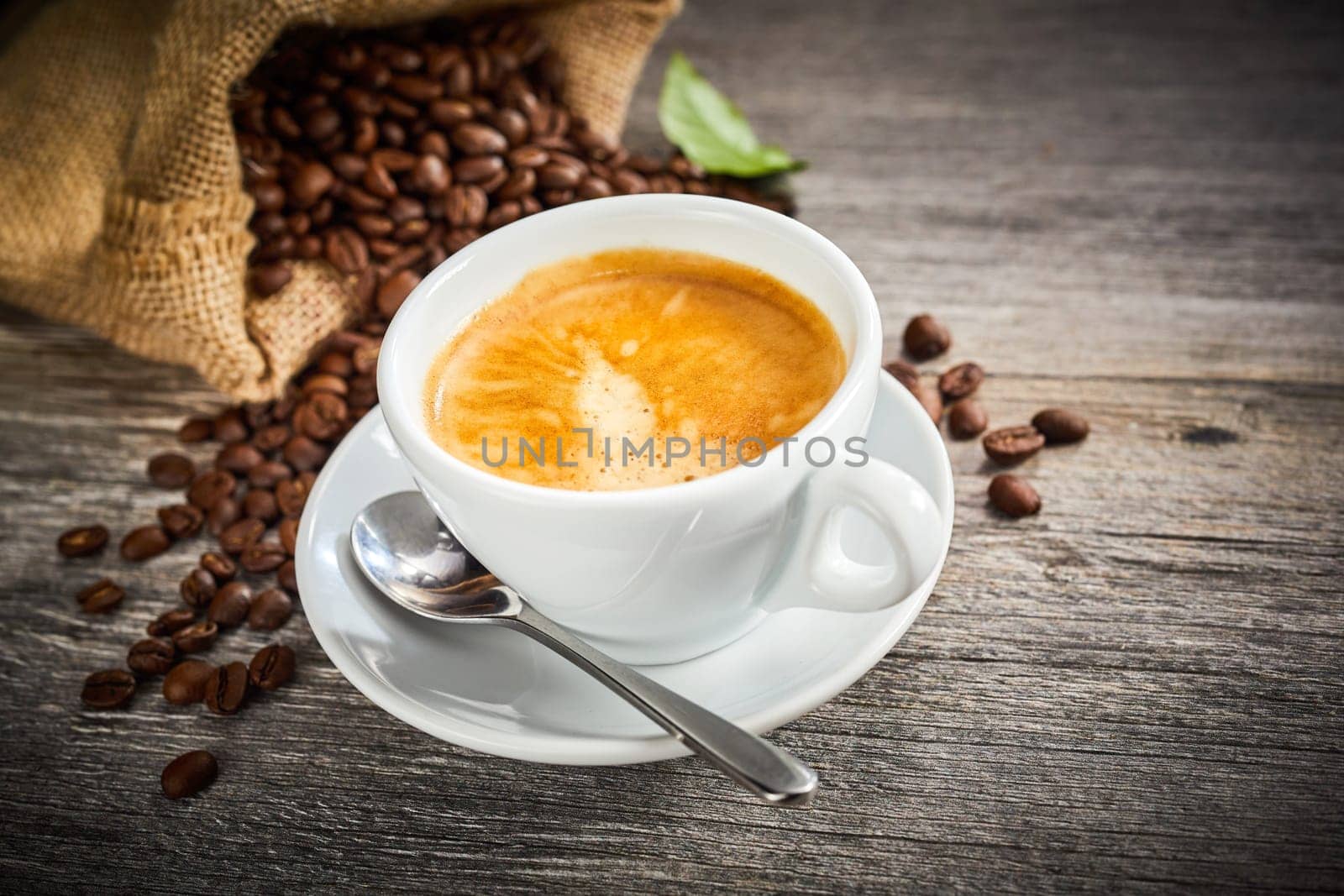 Hot frothy freshly brewed coffee in a white cup and saucer with a bag of roasted beans spilling onto the rustic wood behind and copy space