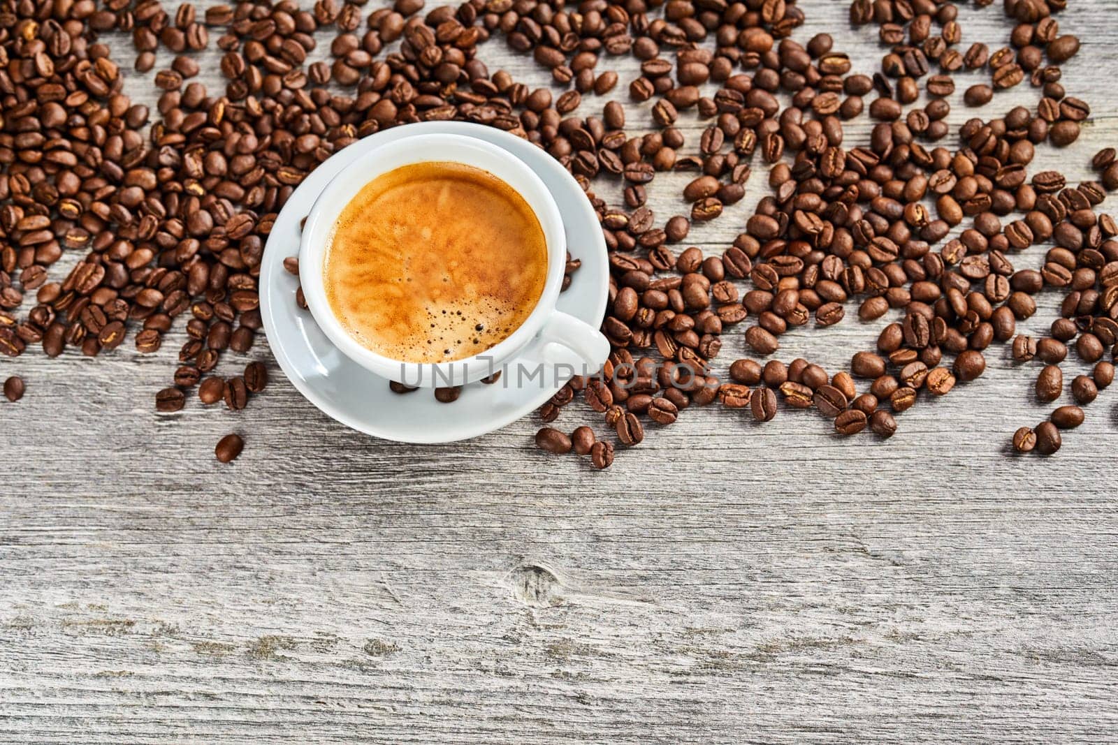 Cup of delicious hot frothy espresso coffee with scattered brown roasted beans viewed top down on rustic weathered wood with copy space below