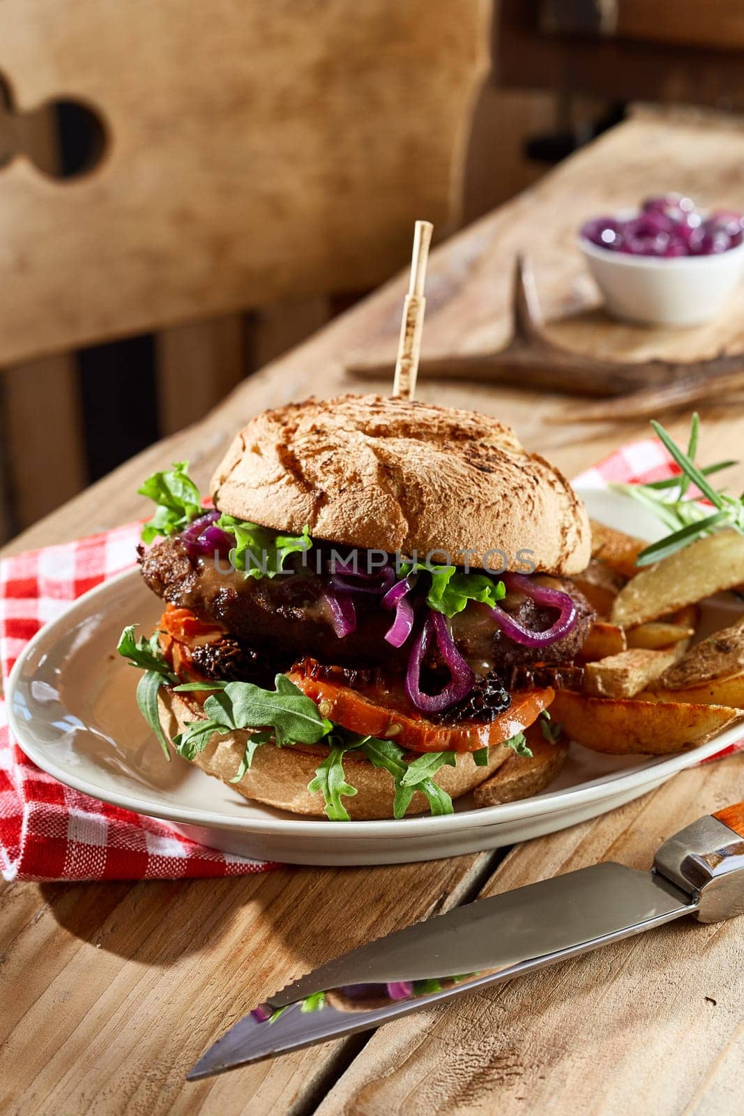 Speciality wild venison burger on a crusty bun trimmed with fresh salad ingredients and served with potato wedges in a close up side view on a rustic table
