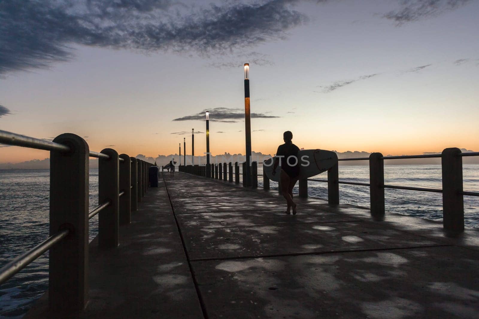 Surfers silhouetted walking beach ocean pier for early morning surfing session.