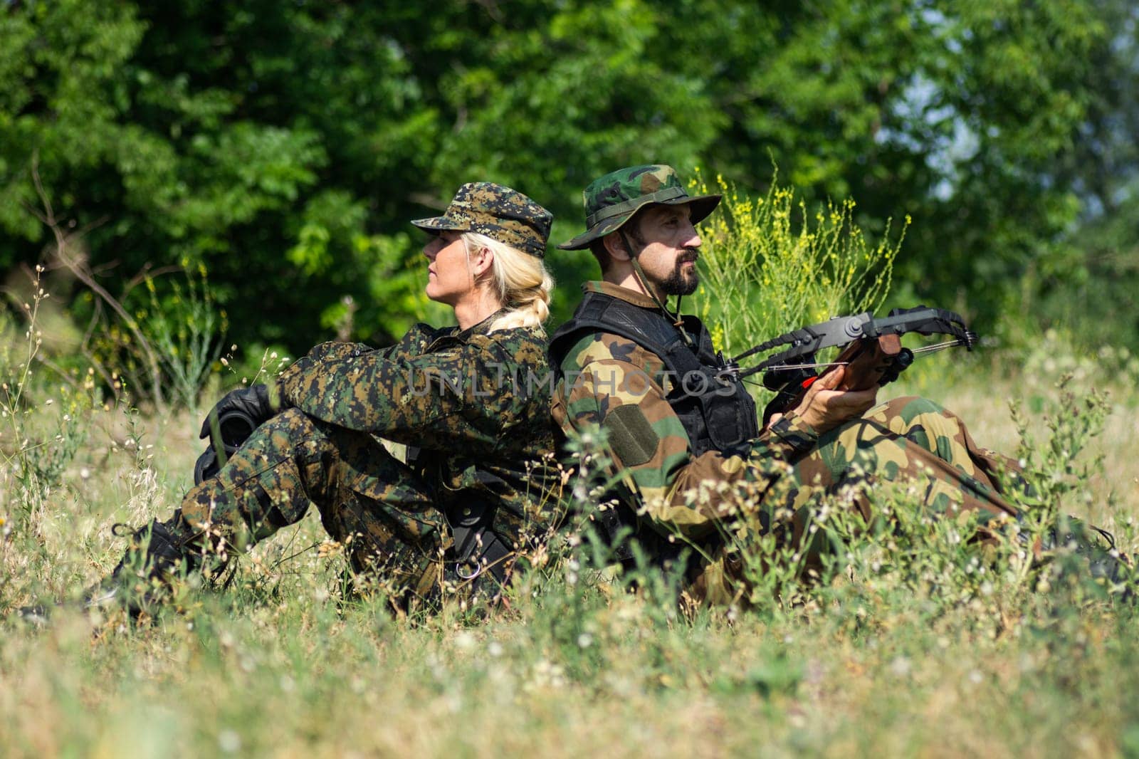 The female and male soldiers in different military uniforms are sitting down in grass in nature.