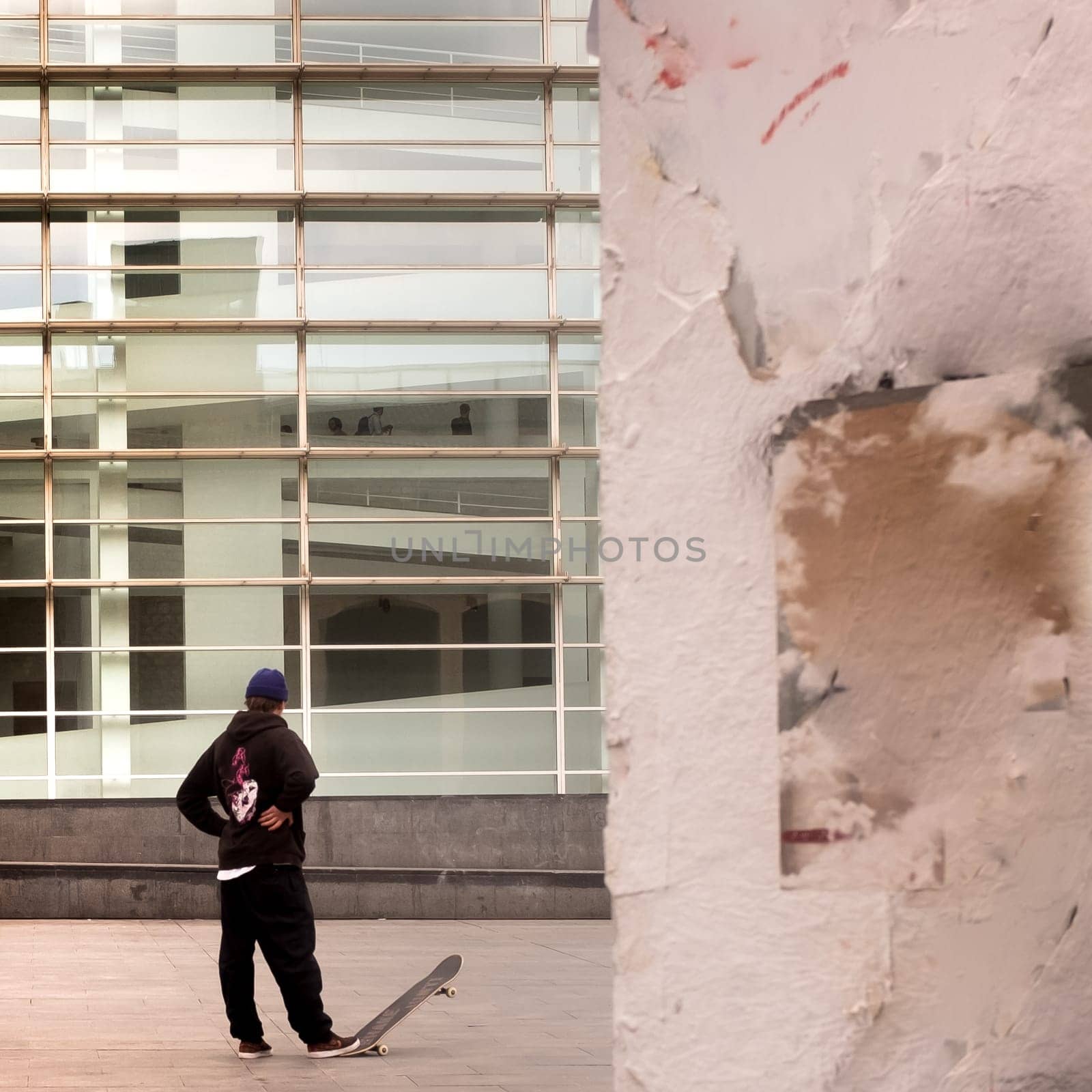 Back view of a young skateboarder standing and relaxed in front of Macba, museum in Barcelona. The boy tipping up the skateboard with his foot. White wall in the foreground on the right side, copy-space.