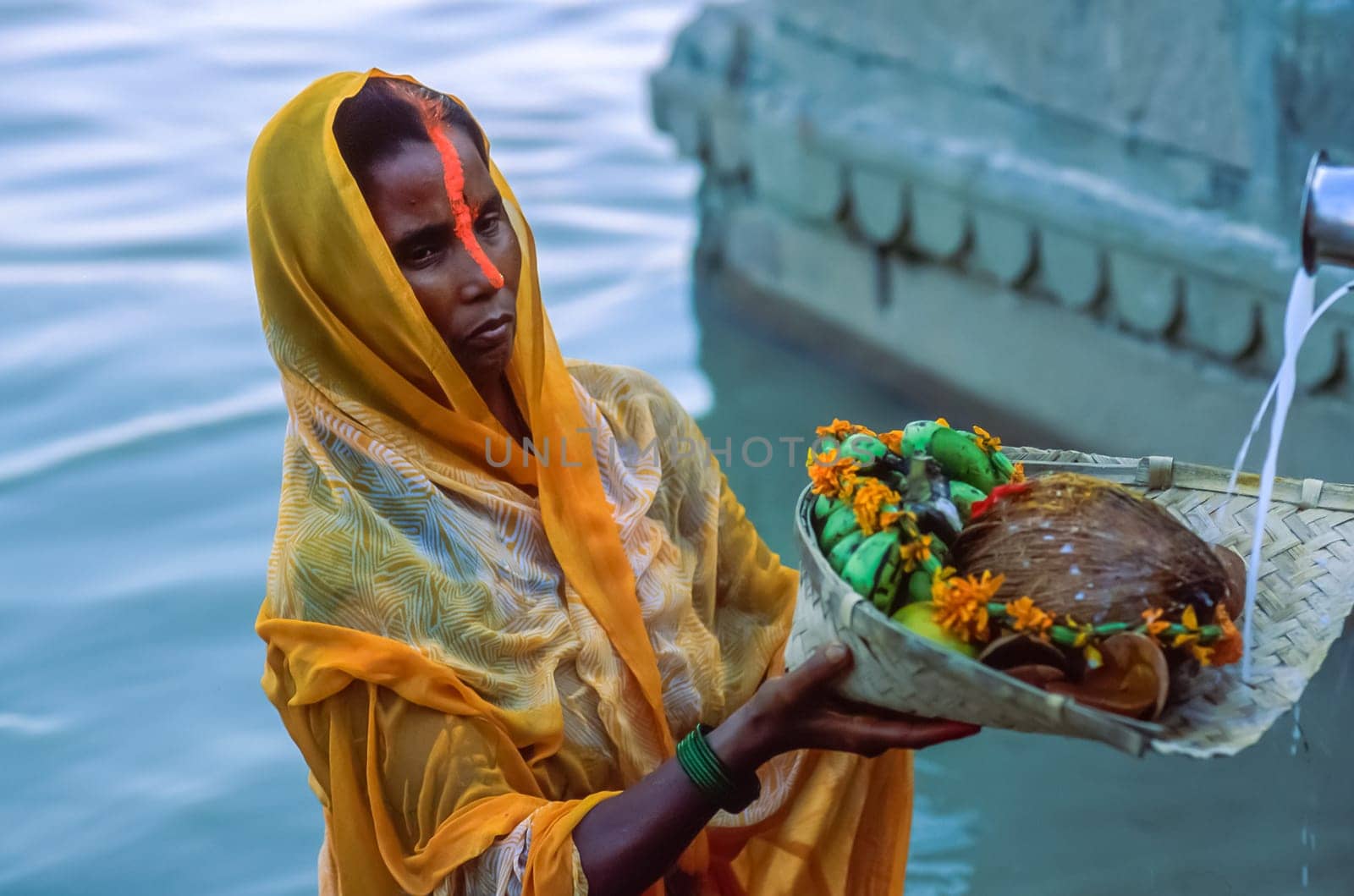 Indian woman devotee wearing a yellow sari standing in the waters of the Ganga River holding in her hands a basket with morning offerings for the Sun God, on the fourth day of the the Chhath Puja celebration, Varanasi, Uttar Pradesh, Northern India.
