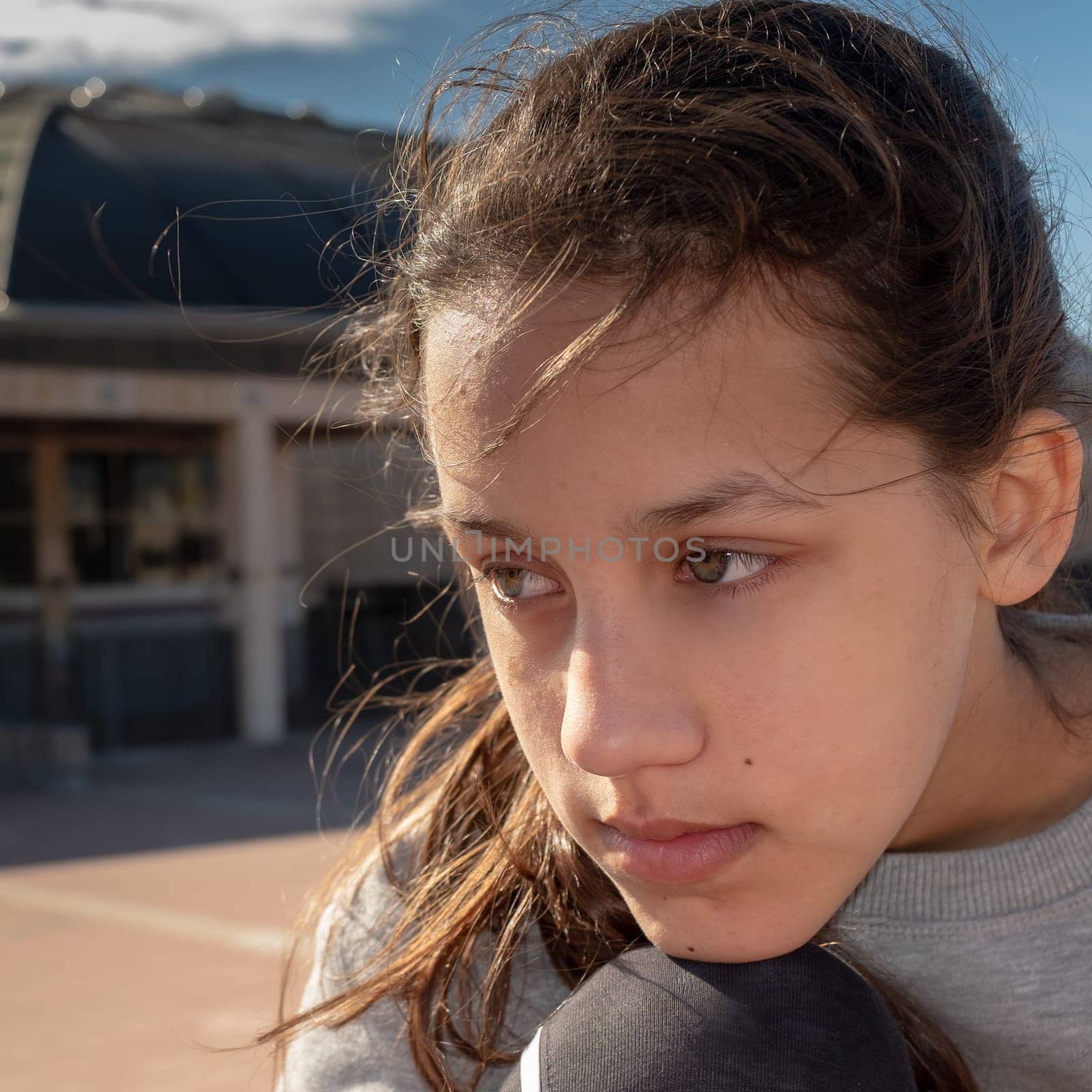 Beautiful teenage girl with long brown hair and cerulean eyes, thoughtful looking elsewhere, sitting relaxed outside, blurred background of a building.