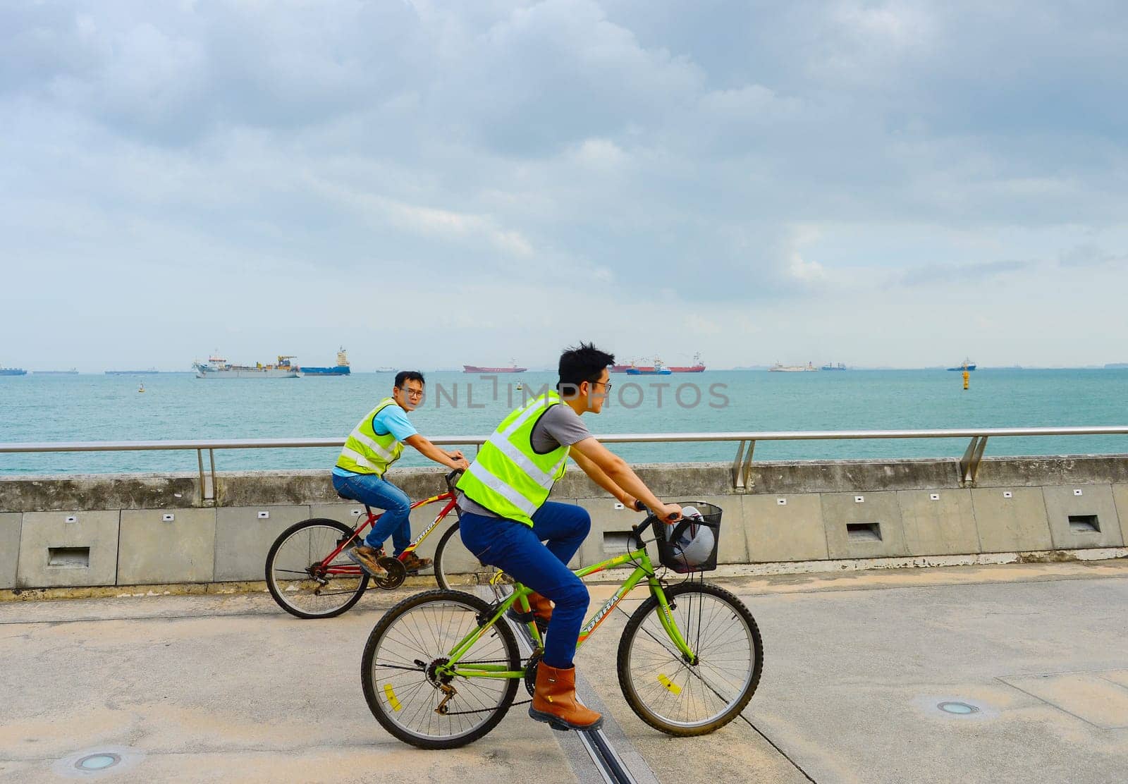 SINGAPORE - FEBRUARY 16, 2107: Two worker in uniform riding the bicycles at Singapore harbor. Industrial cargo ships in the background