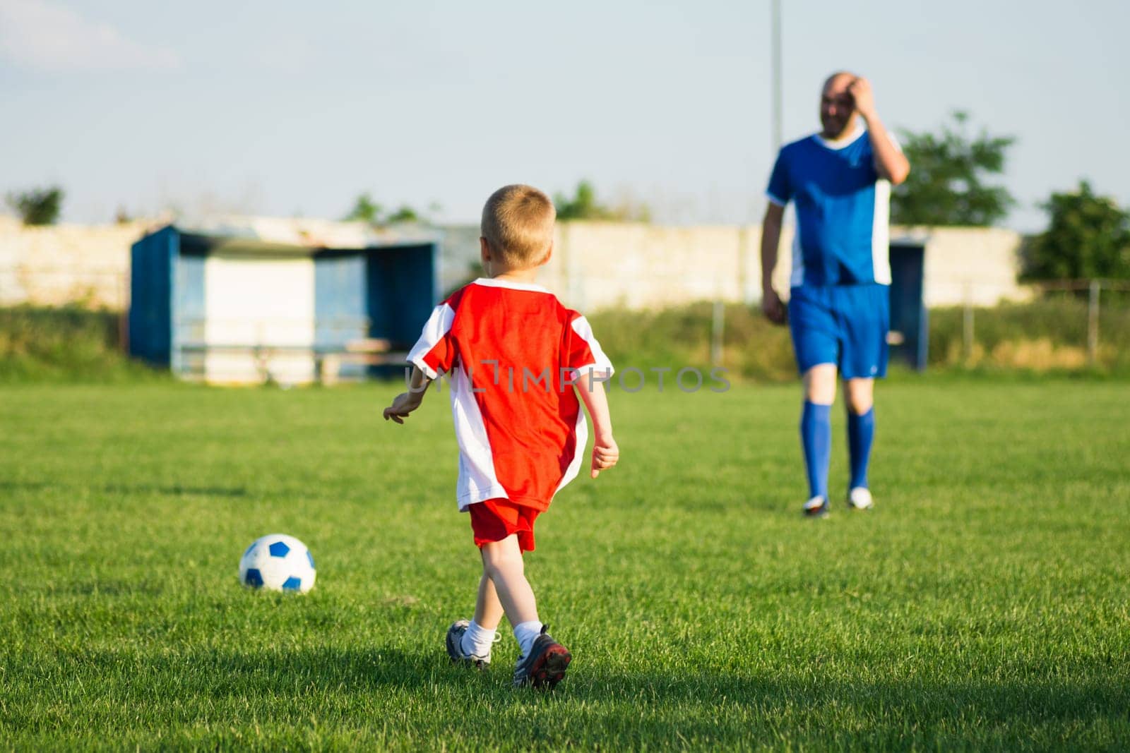 Little soccer boy in red and trainer in blue soccer dress are playing soccer on playing field, soccer training for children concept.