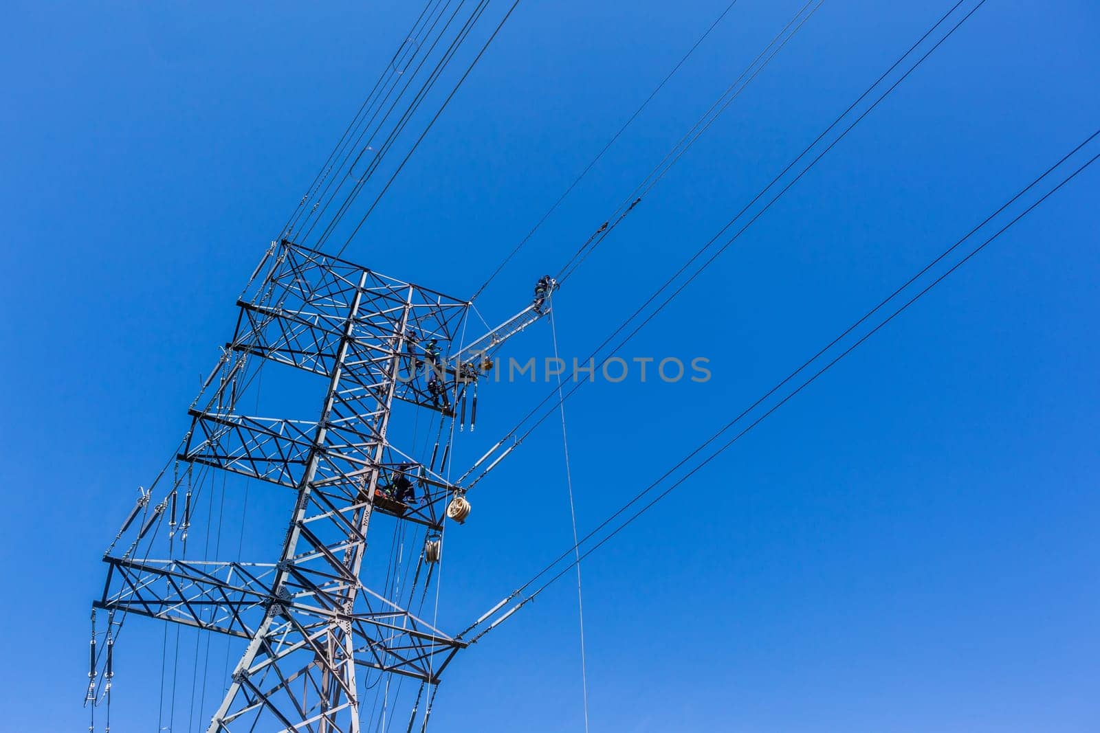 Electricians unidentified men high up suspended on electrical steel tower rigging installing new high voltage electricity power-lines.