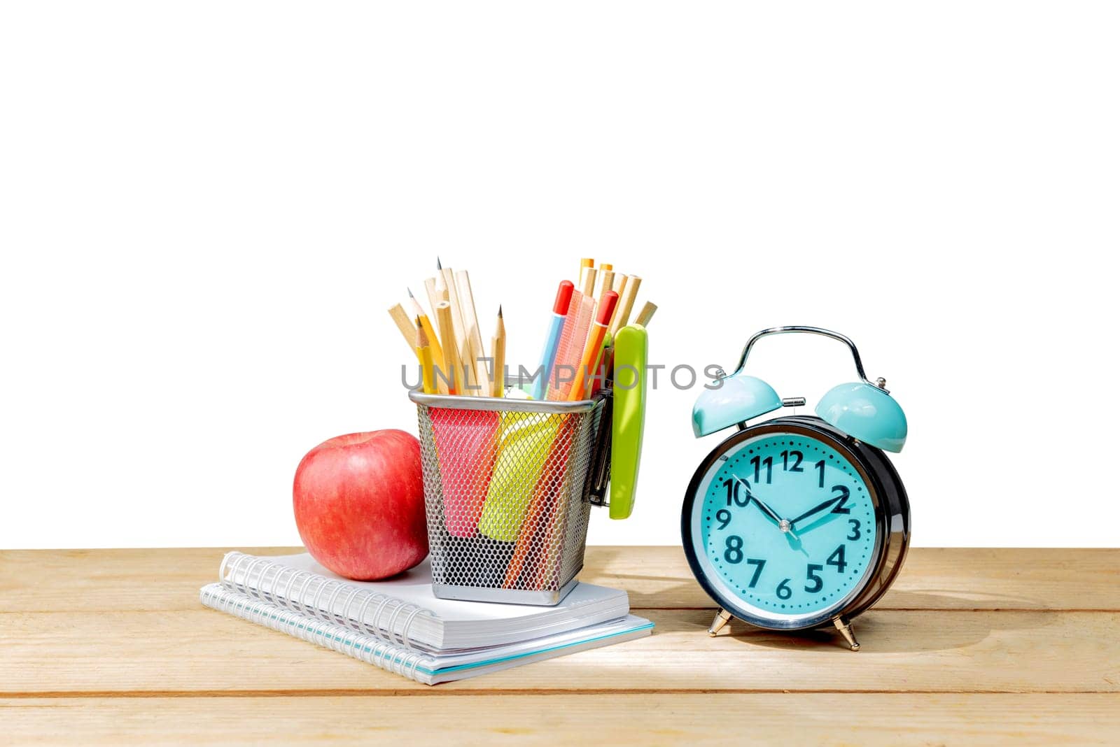 Pile of notebooks with apple and pencils in basket container with green stapler and alarm clock on wooden table isolated over white background. Back to School concept