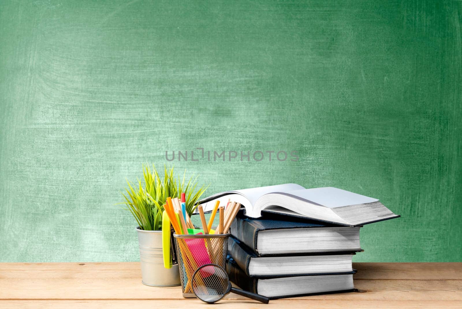 Pile of books with potted plant and pencils in basket container with magnifying glass on the wooden table with chalkboard background. Back to School concept