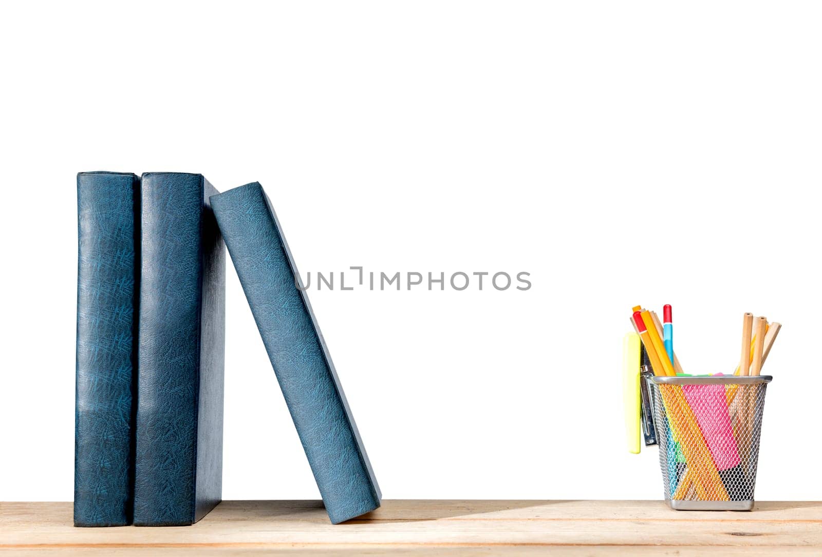 Pile of books standing and pencils in basket container with green stapler on wooden table isolated over white background. Back to School concept