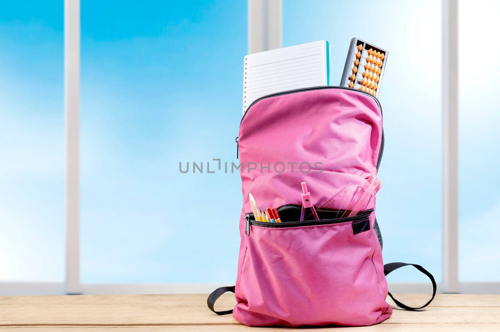 Pink backpack with book and different stationery on the wooden table with window glass background. Back to School concept