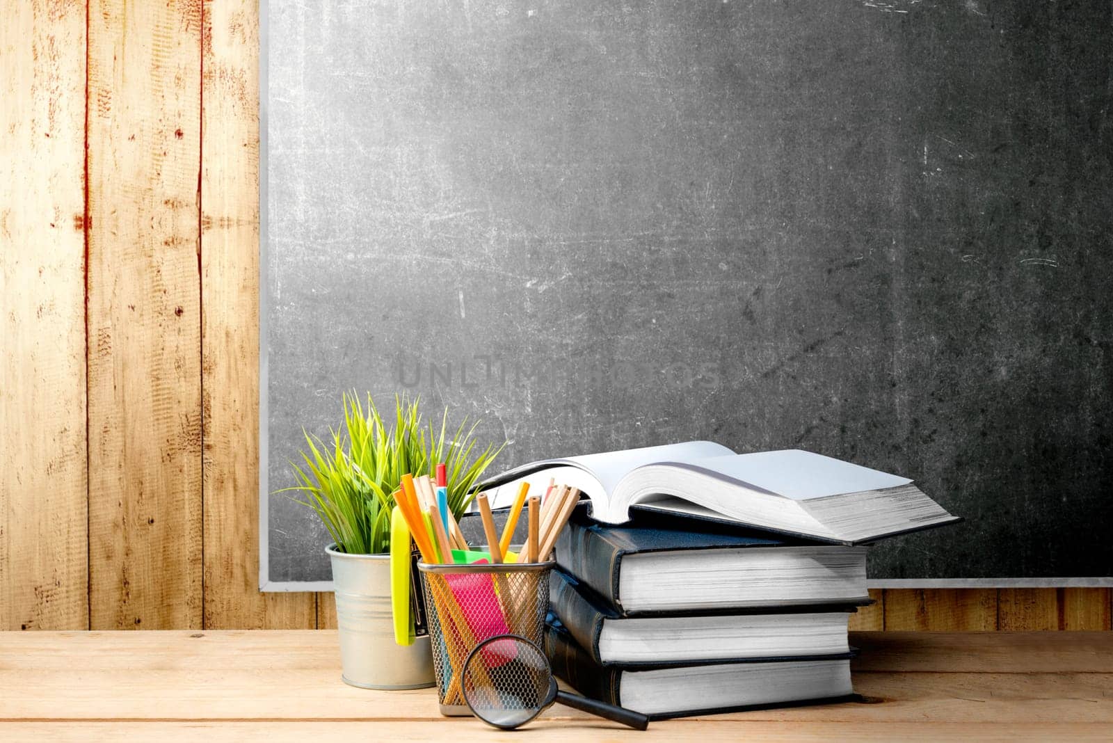 Pile of books with potted plant and pencils in basket container with magnifying glass on the wooden table with blackboard background. Back to School concept