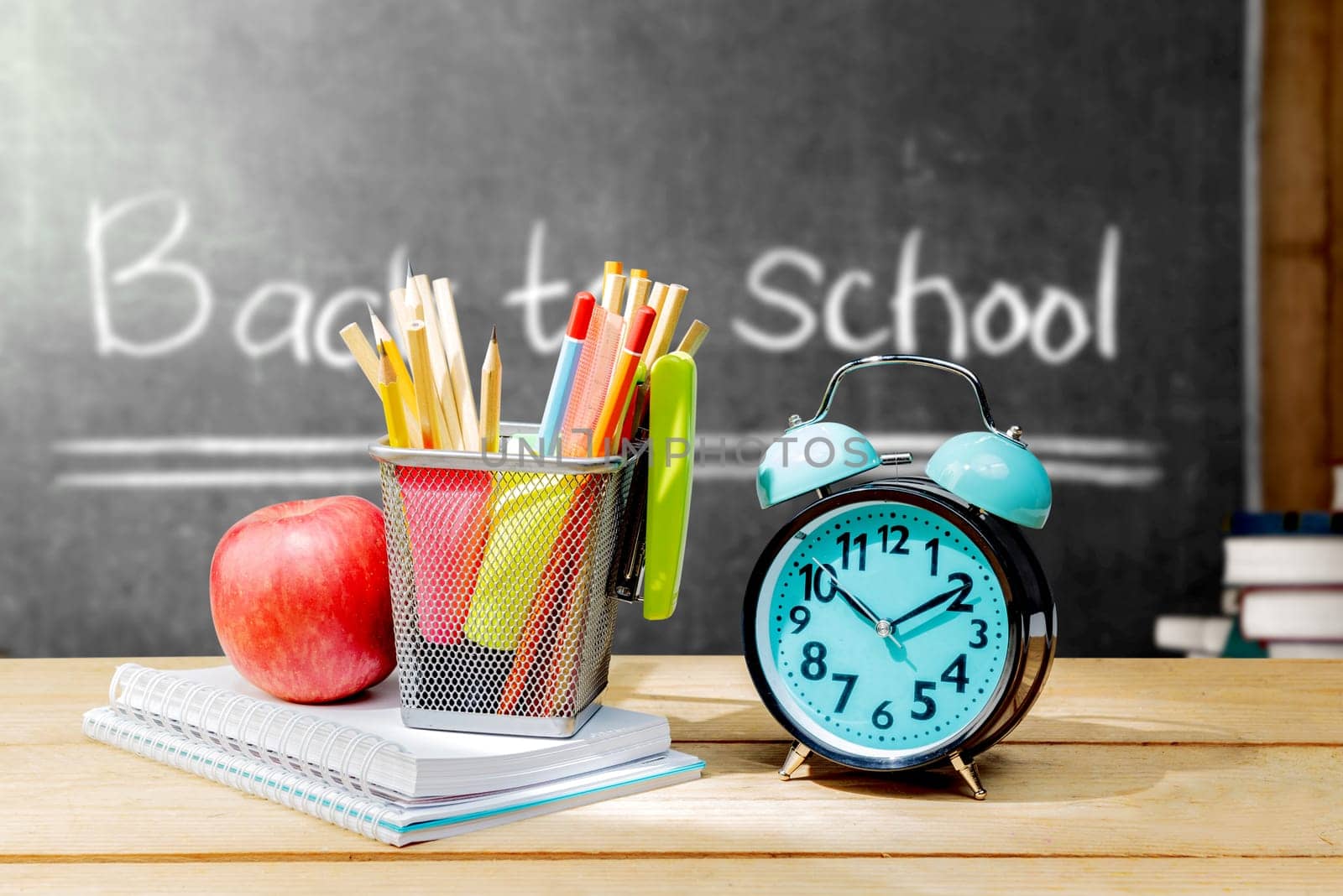 Pile of notebooks with apple and pencils in basket container with green stapler and alarm clock on the wooden table with blackboard background. Back to School concept
