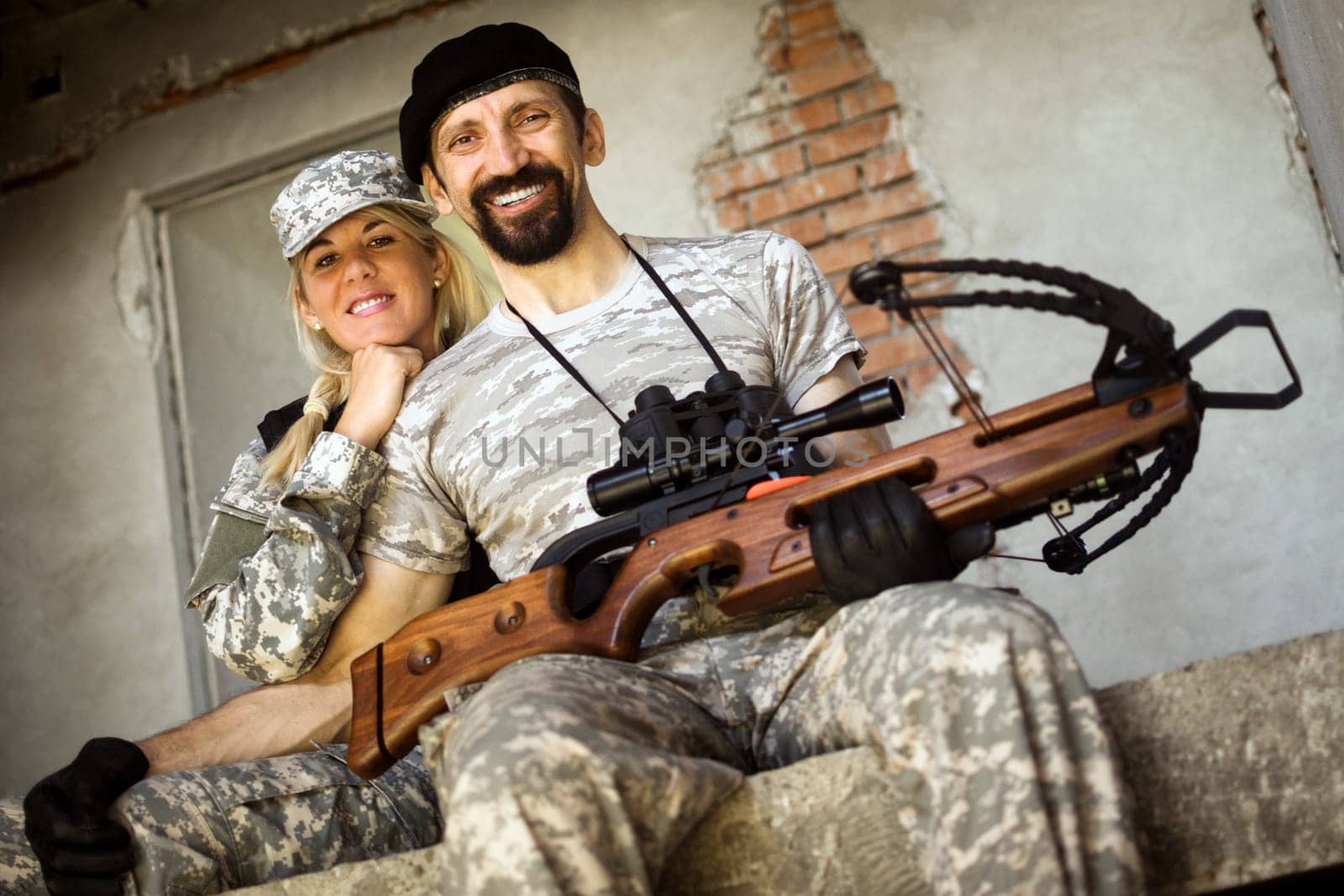 Smiling male and female military persons with crossbow weapon are sitting and looking at camera.