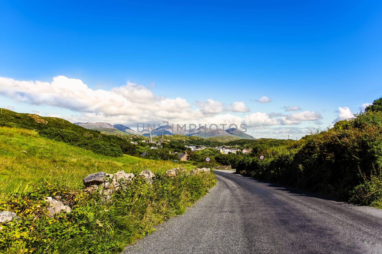 Empty Wild Atlantic Way road between meadows leading in to the village in far distance, blue sky with puffy clouds in golden hour