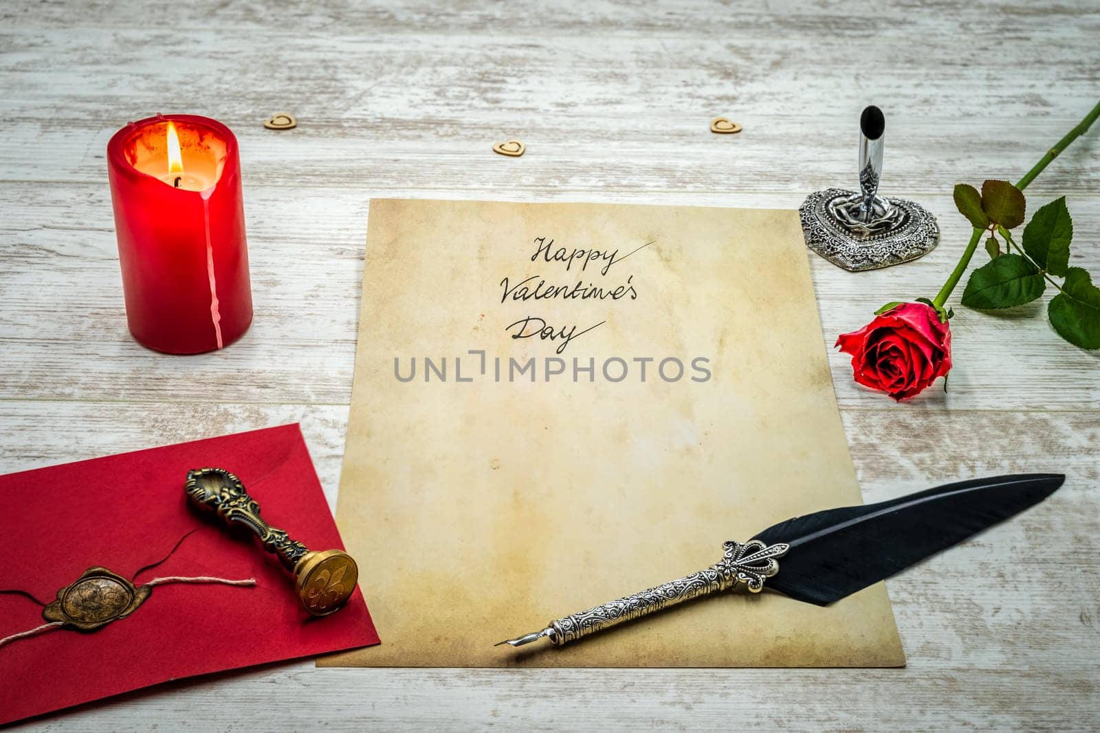 Lovely classic Valentines Day cad, hand written with ink and quill on vintage paper, red envelope with wax seal, red rose, lit candle, silver quill stand on white painted oak - front view