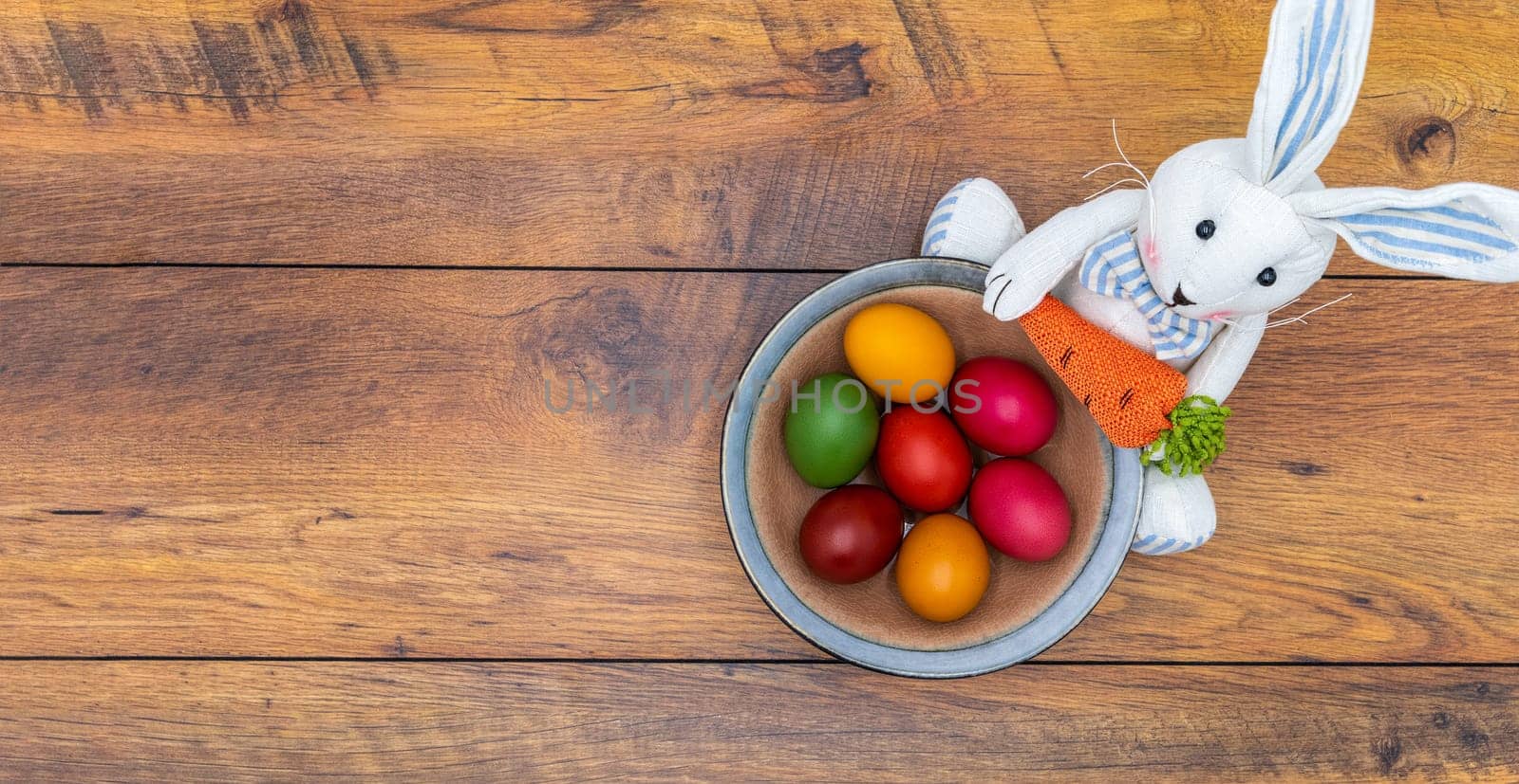 Seven colorful Easter eggs in a brown decorative bowl held by Easter bunny with carrot on vintage oak background, top view