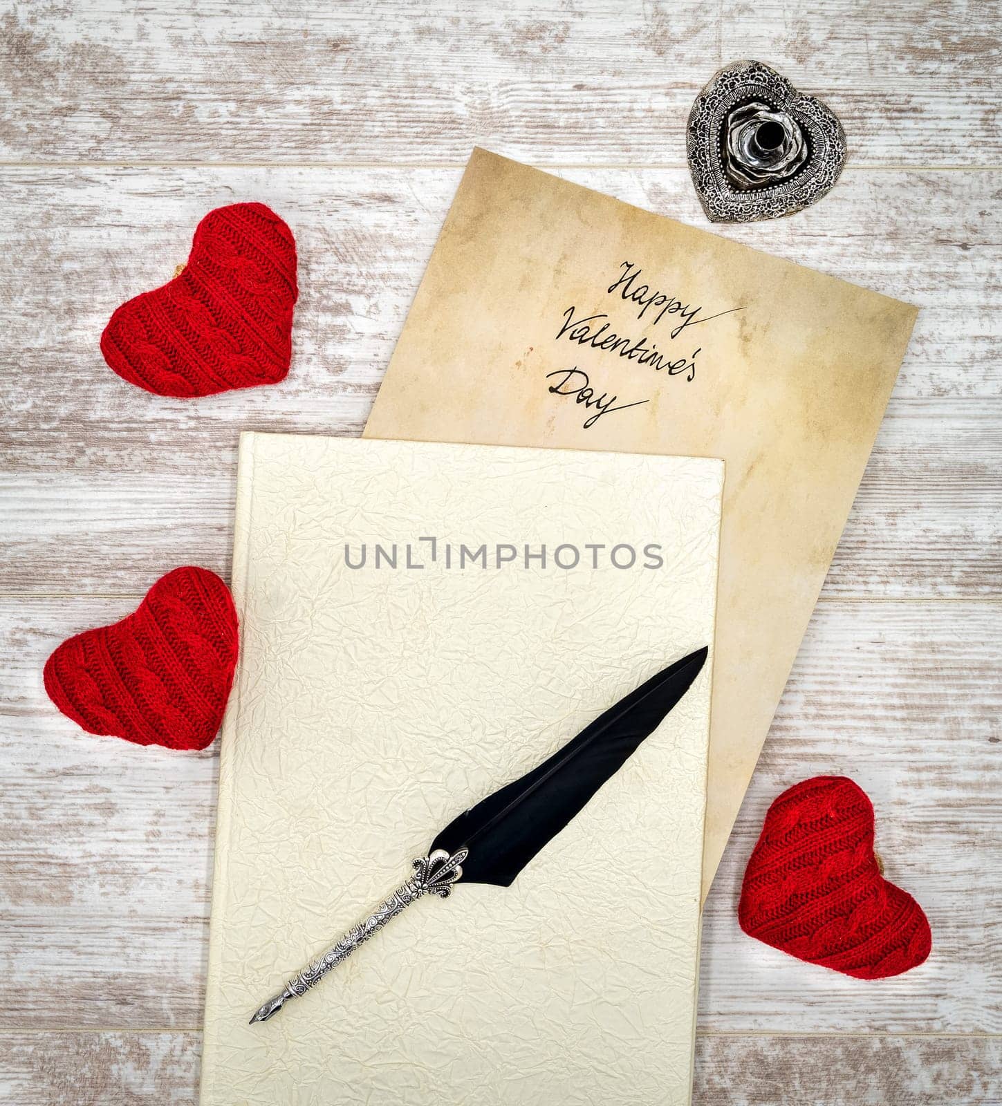 Antique preachment with hand written Happy Valentines Day, sticking out from book, 3 red cuddle heart, detailed silver quill stand in shape of hart, ornamented silver quill on rustic white painted oak