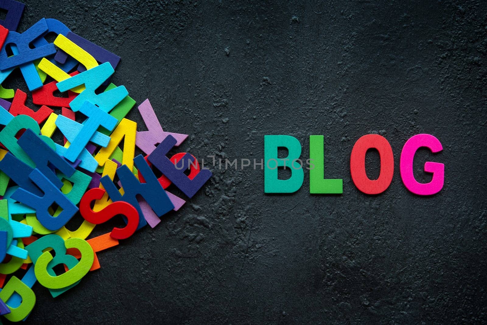 The colorful words "BLOG"" made with wooden letters next to a pile of other letters over dark background."