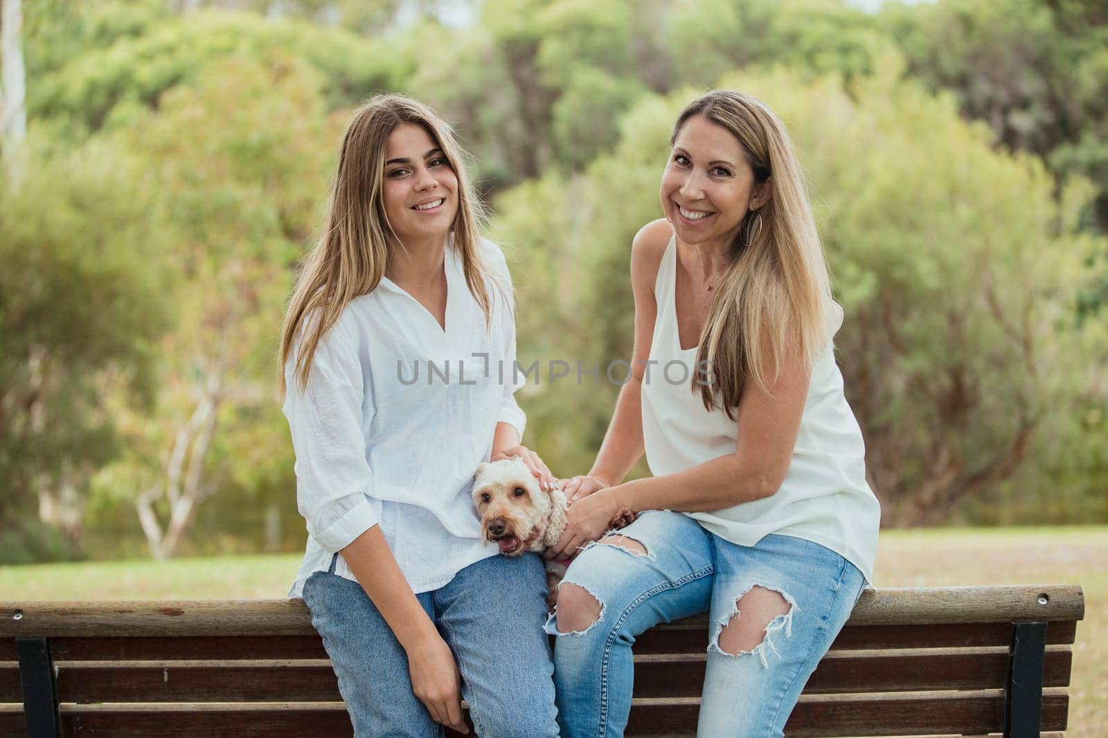 A mother and daughter are sitting by a park bench with their cute dog whilst smiling for a photo.