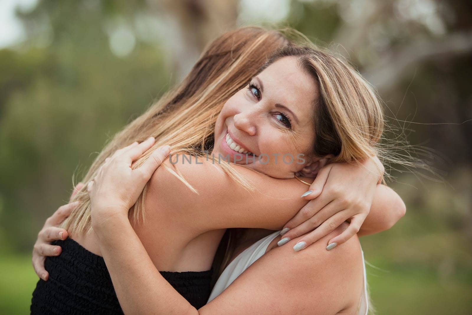A beautiful mother is hugging her daughter outdoors whilst smiling towards the camera.