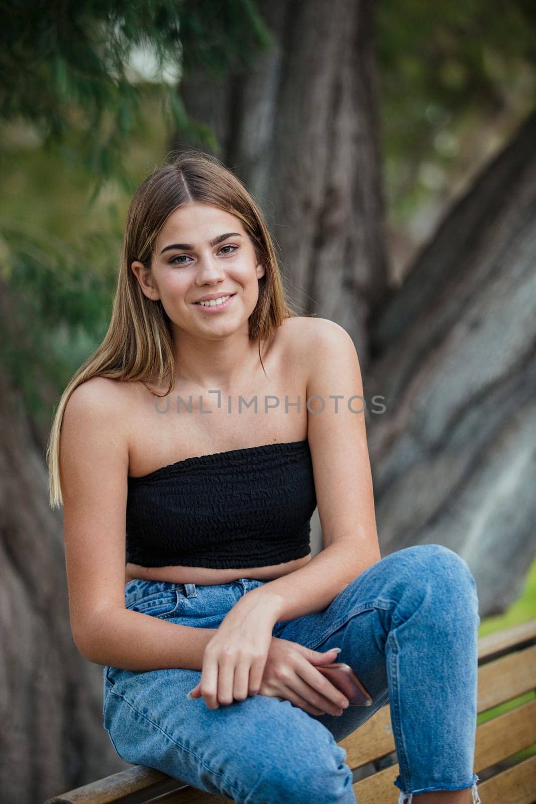 A beautiful teenage girl is sitting on a park bench, smiling and looking towards the camera.