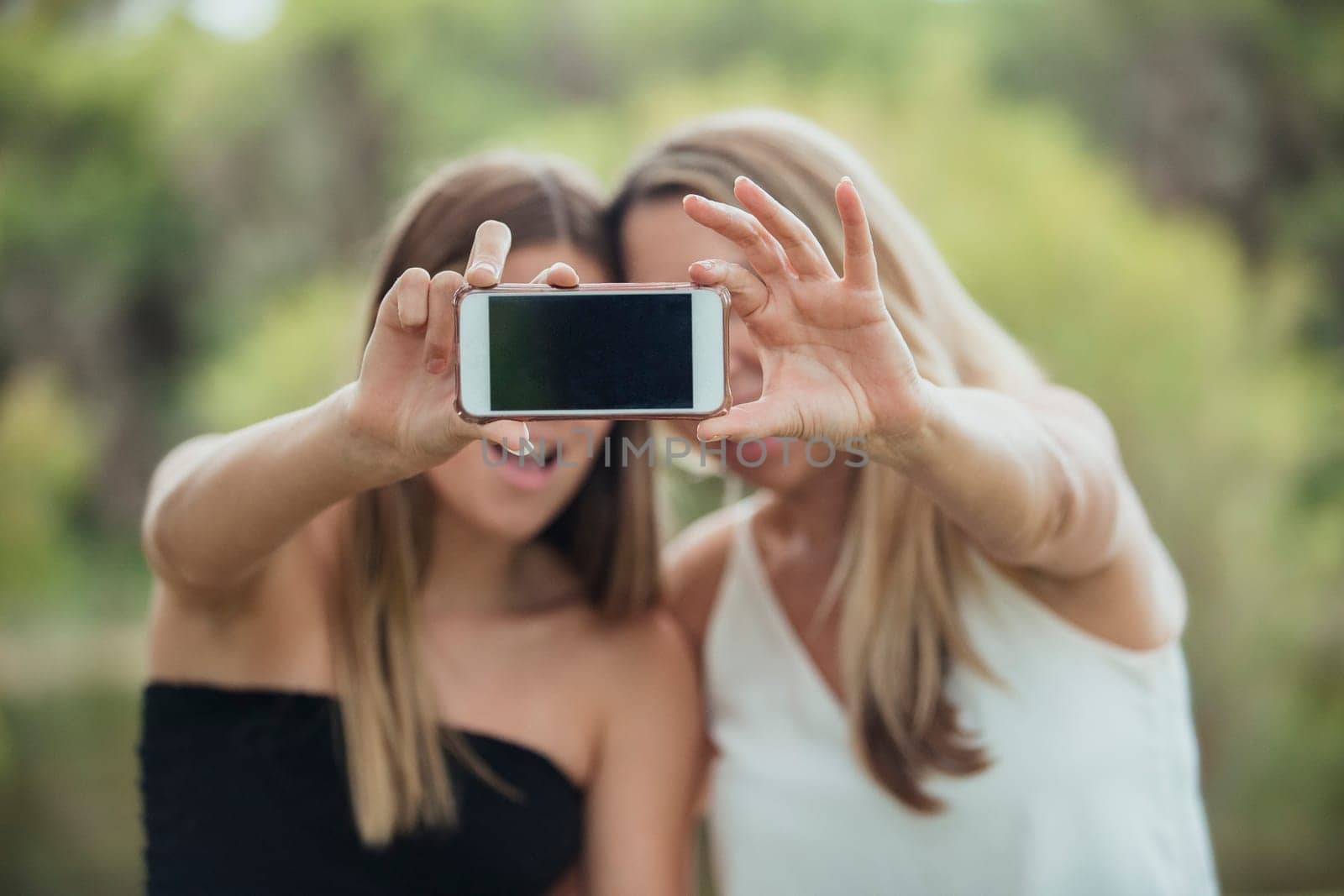 A mother and daughter are standing side by side outdoors capturing a selfie together.