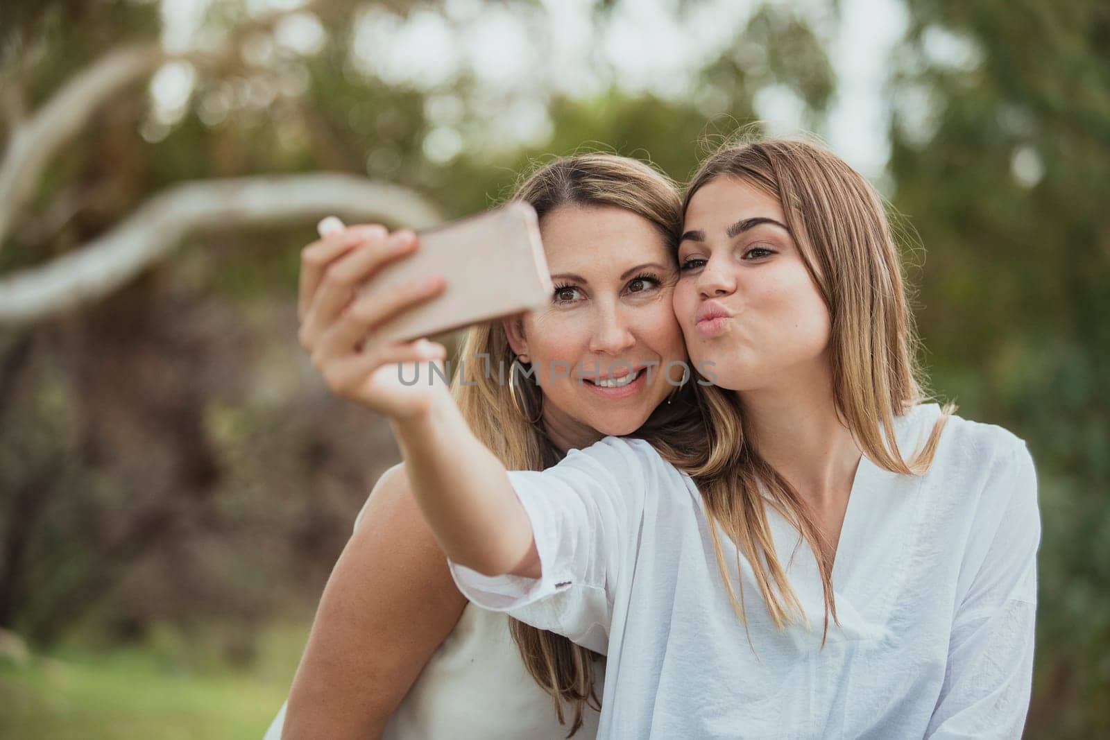 A mother and daughter are side by side in the park posing for a selfie with their dog.