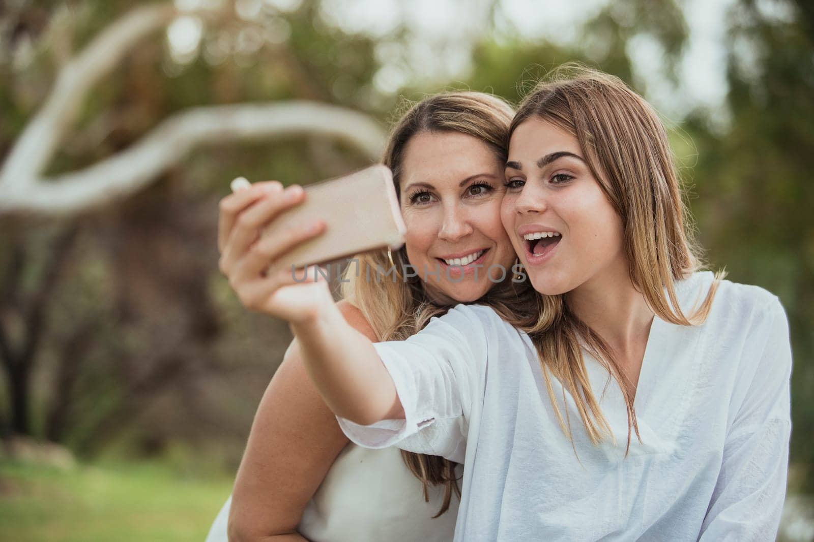 A mother and daughter are side by side in the park posing for a selfie with their dog.
