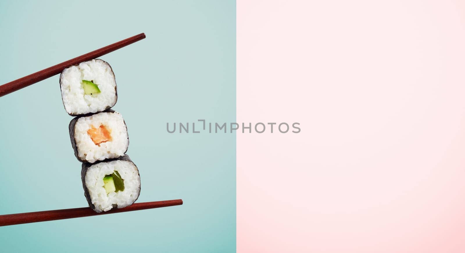 Chopsticks holding three seaweed wraps with rice and avocado over blue half of two tone background. Includes copy space.
