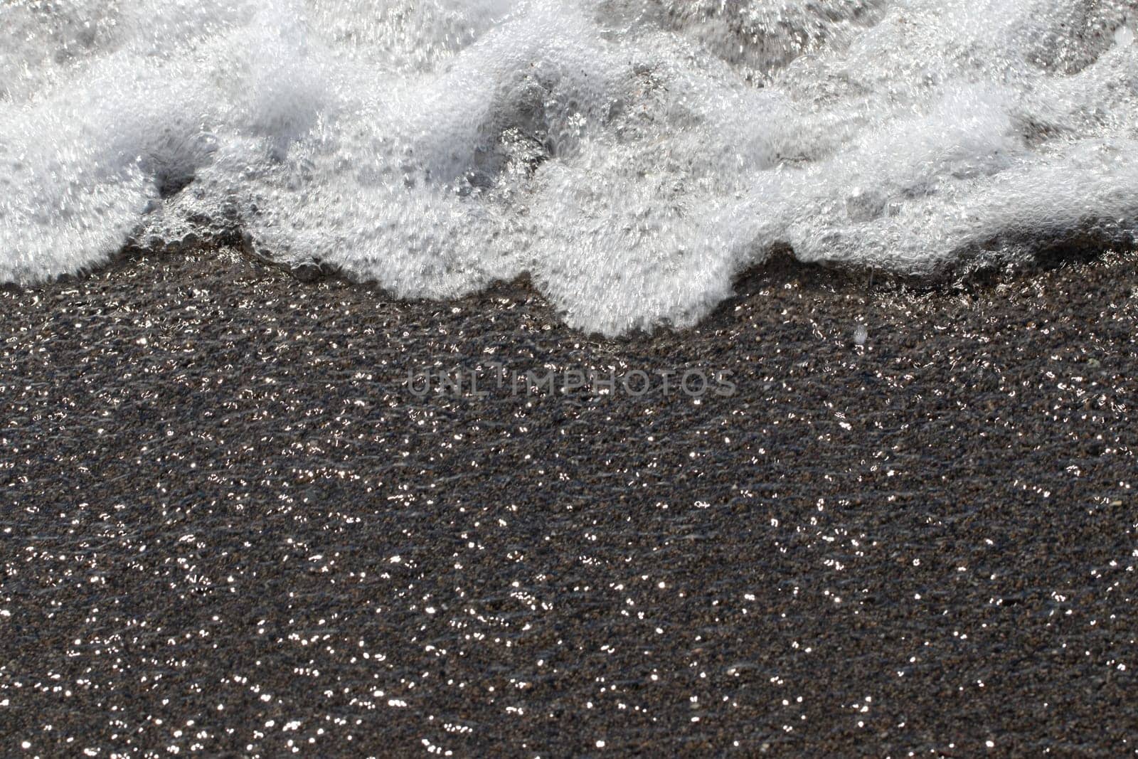 Wave foaming on the sand. Closeup of sea waves with white foam on the sandy shore with sun reflections effect.