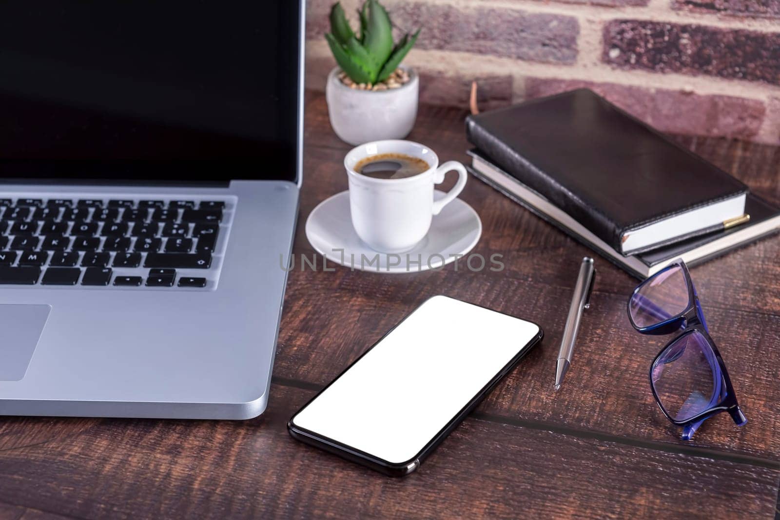 Laptop notebook with blank screen and cup of coffee and notepad pen and books and smartphone on wooden table. .Mock up on wooden table with laptop with blank screen, cup of coffee, pen, books and flower..Focus on smart-phone and glasses.