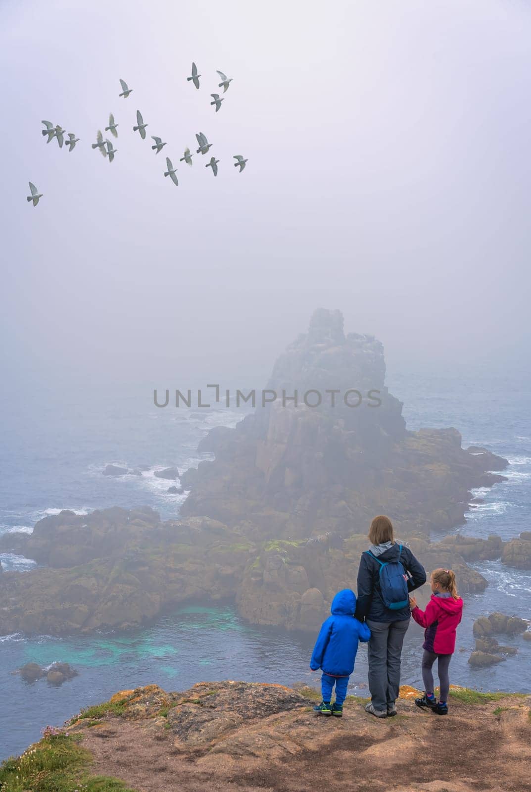 Mother with her children standing on the top of a cliff looking at the rock in the sea at the Lands End - the most westerly point of England, Penwith peninsula, Cornwall