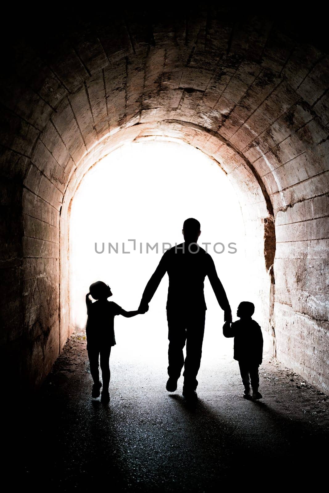Father walking with his two little siblings children, boy and girl, towards the light at the end of a long dark tunnel