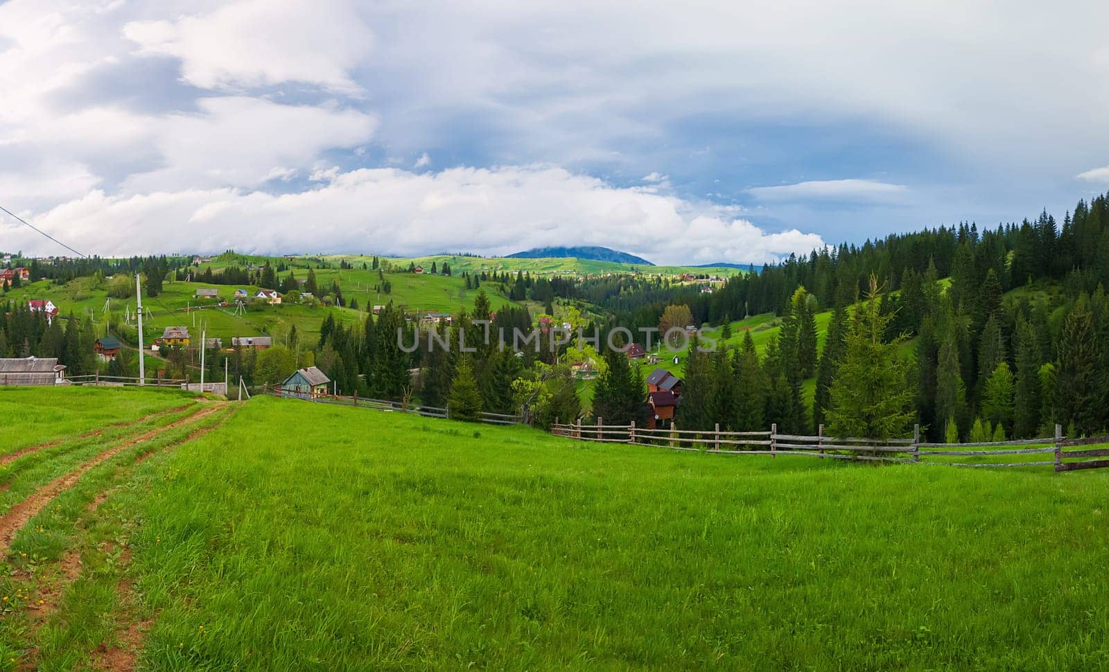 Picturesque spring mountains scene with wooden split rail fence across a green and lush pasture, a country road and old houses on the valley surrounded by pine woods. Yablunytsya, Ukraine.