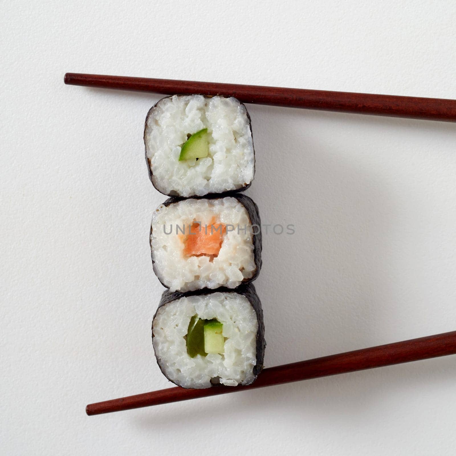Food styling concept with three maki sushi rolls with nori seaweed, salmon and avocado pear placed between bamboo chopsticks on white with copy space