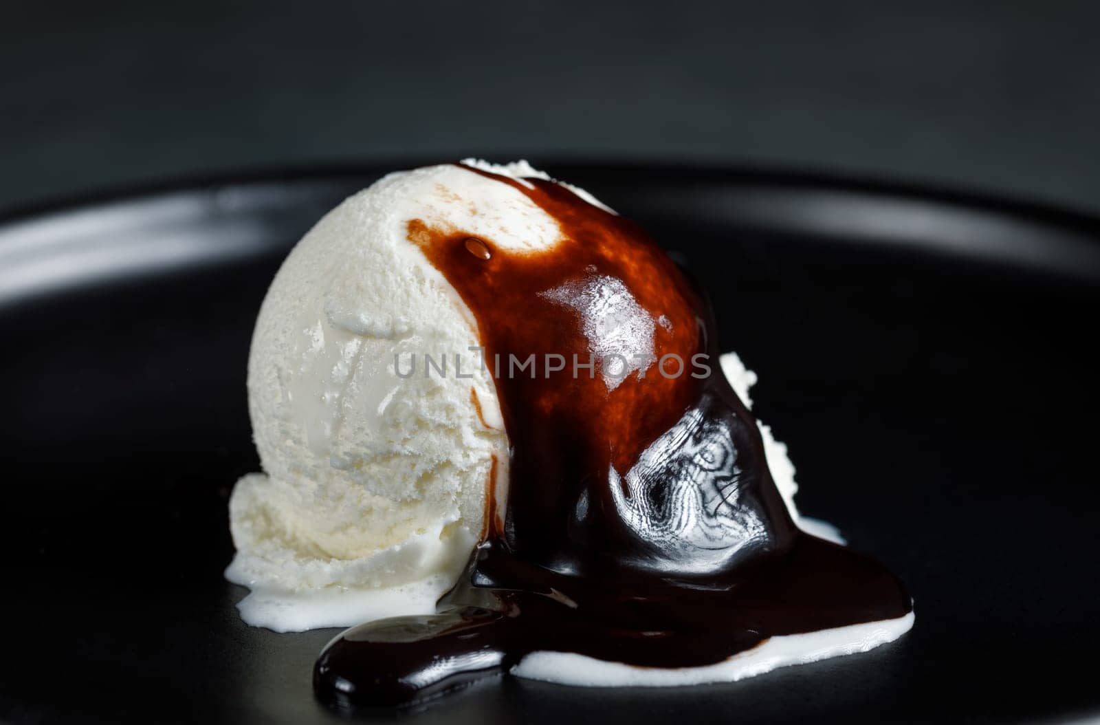 White cream ball on a black ceramic dish on a dark background, poured with chocolate sauce
