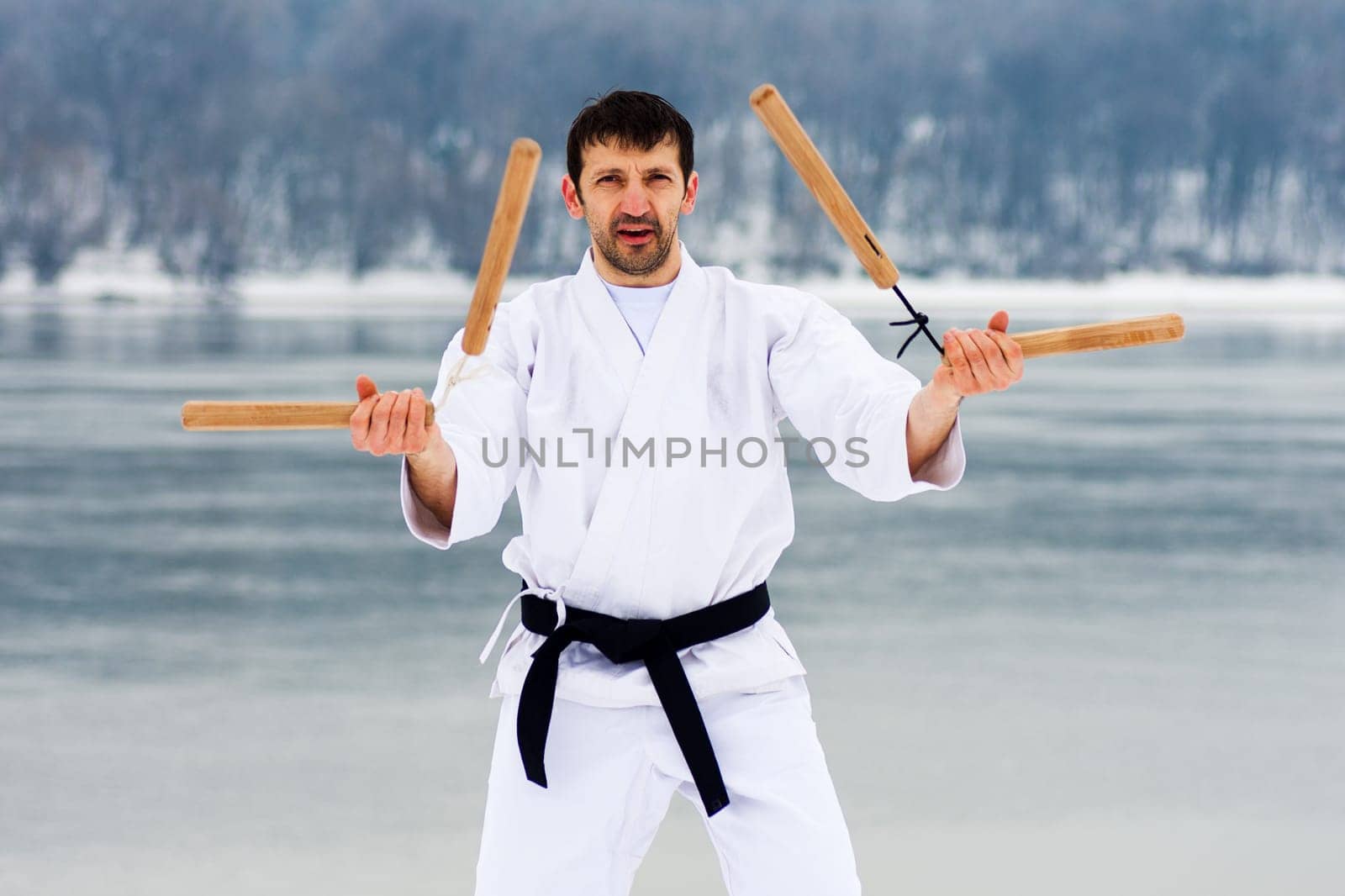 The middle aged man in white kimono and black belt is practicing martial arts with two wooden nunchaku at winter.