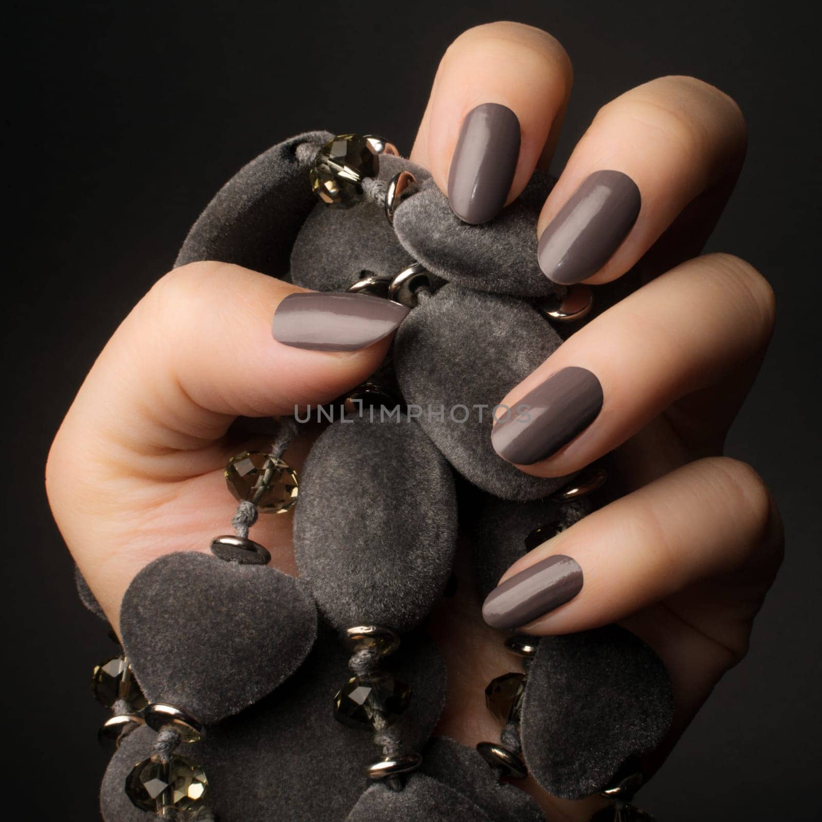 Female hand with shiny dark gray nails is holding a gray jewelry on dark gray background.
