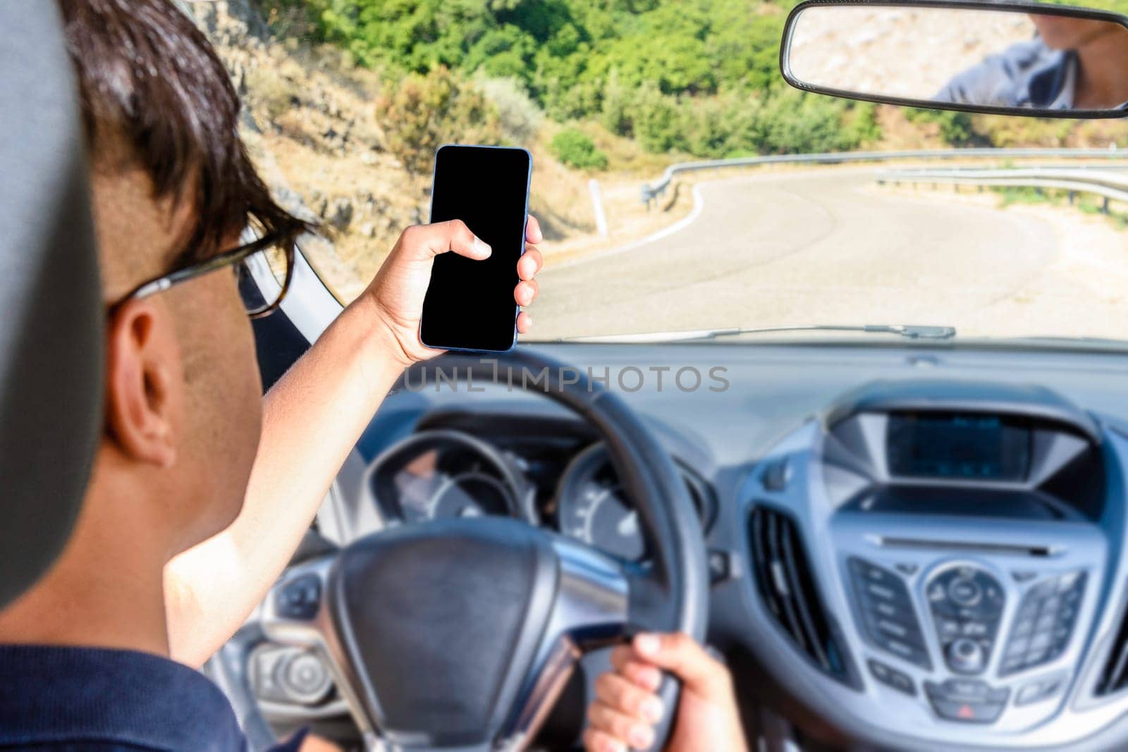 Young driver, using smartphone, on the road in the car. Distraction and danger on the road. Smartphone blank area and free background.