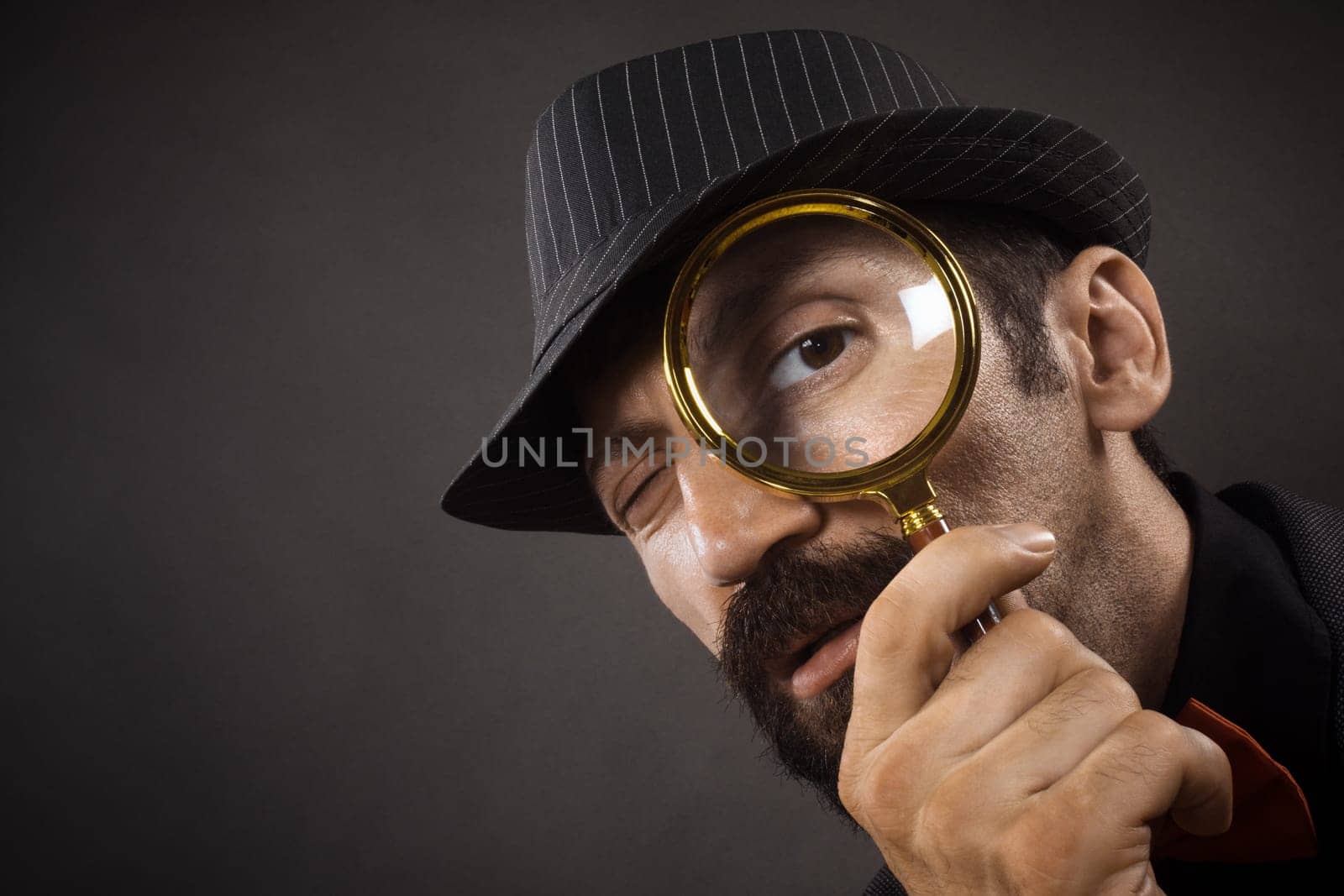 The old-fashioned detective with hat is looking through loupe or magnifier on dark gray background.