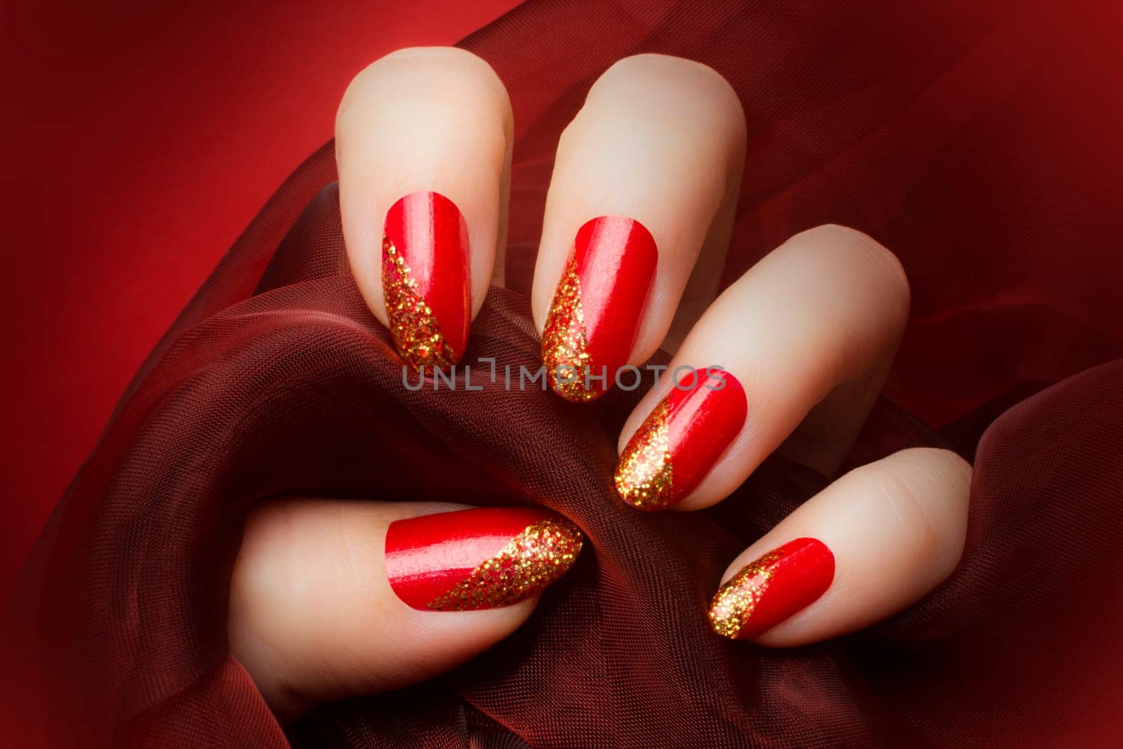 Female hand with beautiful red and golden glittered nails is holding a dark red textile on red background, manicure and nail care concept.