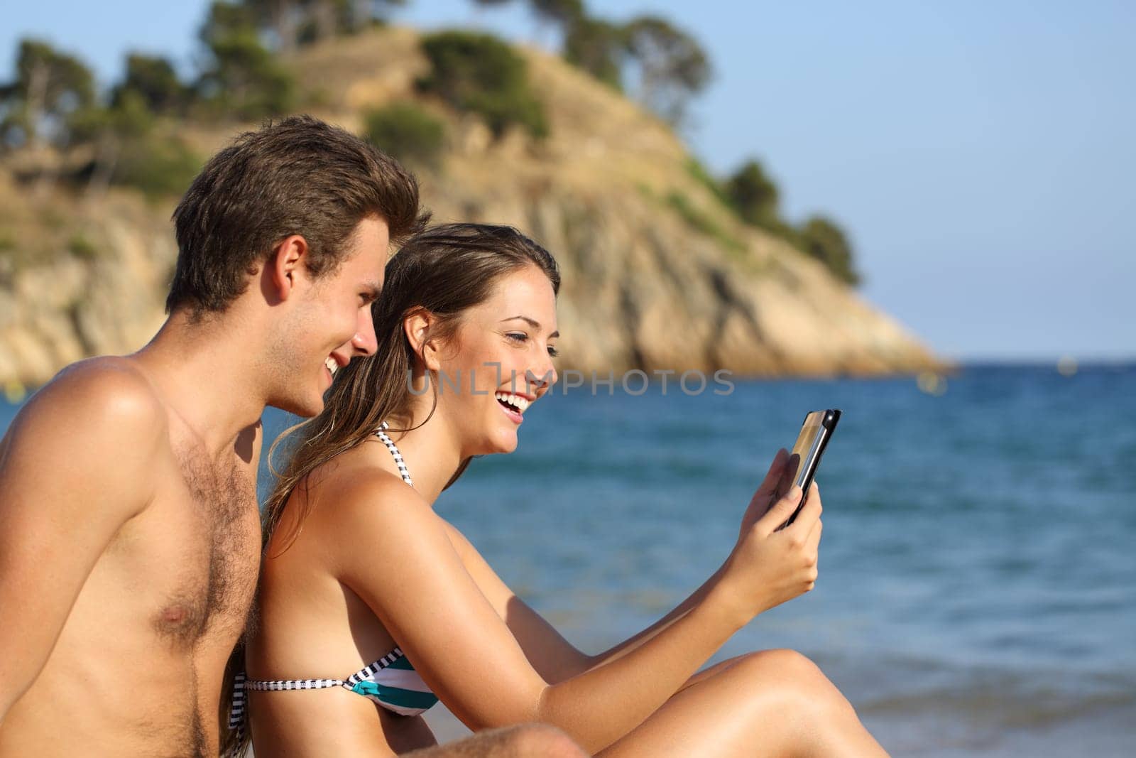Side view portrait of a happy couple on the beach checking tablet on summer vacation